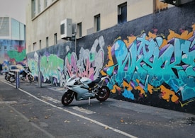 A street scene featuring a line of parked motorcycles against a building wall adorned with vibrant graffiti. The graffiti displays a variety of colors and styles, creating a striking urban artwork. The street surface is paved and there are scattered leaves around.