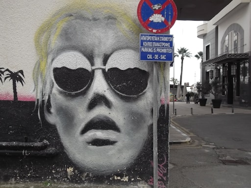 A street art mural on a wall depicts a stylized face wearing large circular sunglasses, painted in black and white tones. The face is positioned next to a realistic street scene featuring a 'No Parking' sign with text in both English and Greek. The background includes hints of trees and urban architecture.