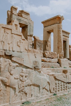 Ancient stone carvings and ruins featuring detailed reliefs of figures and mythological creatures. The structures have ornate columns and a weathered appearance, reflecting historical architecture.