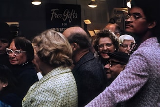 A diverse group of people gathered, seemingly engaged in conversation or waiting for something. They are positioned closely, creating a sense of crowding. Most individuals appear to be older adults, and some are wearing glasses. The background features a sign that says 'Free Gifts for New Accounts.' The lighting is subdued, suggesting an indoor setting or an overcast day.