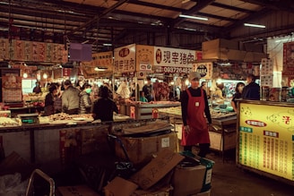 A bustling indoor market with various stalls selling meats and produce. People are seen shopping and interacting, with vendors wearing aprons and some wearing masks. The market is dimly lit with warm, ambient lighting and signs in Asian writing hanging above the stalls.