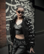 A person is dressed in fashionable, dark clothing, including a shiny black crop top and loose pants. They are wearing sunglasses and leaning against a railing. The background features a wall with a stylized, artistic pattern in black and white.