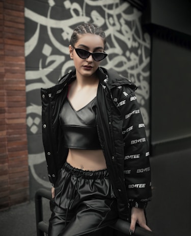 A person is dressed in fashionable, dark clothing, including a shiny black crop top and loose pants. They are wearing sunglasses and leaning against a railing. The background features a wall with a stylized, artistic pattern in black and white.