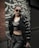A person is dressed in fashionable, dark clothing, including a shiny black crop top and loose pants. They are wearing sunglasses and leaning against a railing. The background features a wall with a stylized, artistic pattern in black and white.