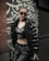 A person is dressed in fashionable, dark clothing, including a shiny black crop top and loose pants. They are wearing sunglasses and leaning against a railing. The background features a wall with a stylized, artistic pattern in black and white.