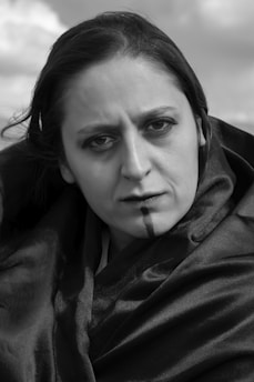 A person with a serious expression is wrapped in a dark, silky fabric. The background is blurred, featuring clouds, creating a dramatic and introspective scene. The black and white color scheme accentuates the mood.