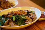 A shallow white bowl contains a savory dish with thin slices of cooked beef and vibrant green leafy vegetables, garnished with crispy fried garlic pieces. A separate dish with red sauce is placed near the bowl, all set on a wooden table surface.