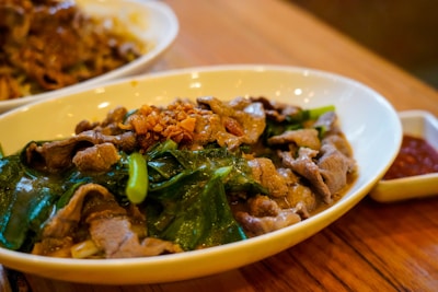 A shallow white bowl contains a savory dish with thin slices of cooked beef and vibrant green leafy vegetables, garnished with crispy fried garlic pieces. A separate dish with red sauce is placed near the bowl, all set on a wooden table surface.