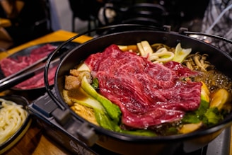 A steaming hot pot dish filled with various ingredients, including thinly sliced red marbled beef, leafy greens, mushrooms, tofu, and noodles, is presented in a black metal pot. The dish appears richly colored and well-cooked, suggesting a savory and hearty meal.
