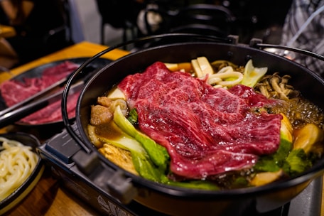 A steaming hot pot dish filled with various ingredients, including thinly sliced red marbled beef, leafy greens, mushrooms, tofu, and noodles, is presented in a black metal pot. The dish appears richly colored and well-cooked, suggesting a savory and hearty meal.