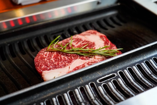 A raw marbled steak is placed on a grill with a sprig of rosemary on top. The grill has visible metal grates and appears to be hot, ready for cooking the meat. The marbling in the steak suggests it's a high-quality cut.