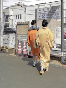 Two individuals wearing traditional kimonos walk along a street. They are moving towards a sign that provides directions to Nezu-jinja Shrine and Nippon Medical School Hospital. The scene includes several traffic cones and a planter with greenery. The background features buildings and overhead wires, contributing to an urban setting.
