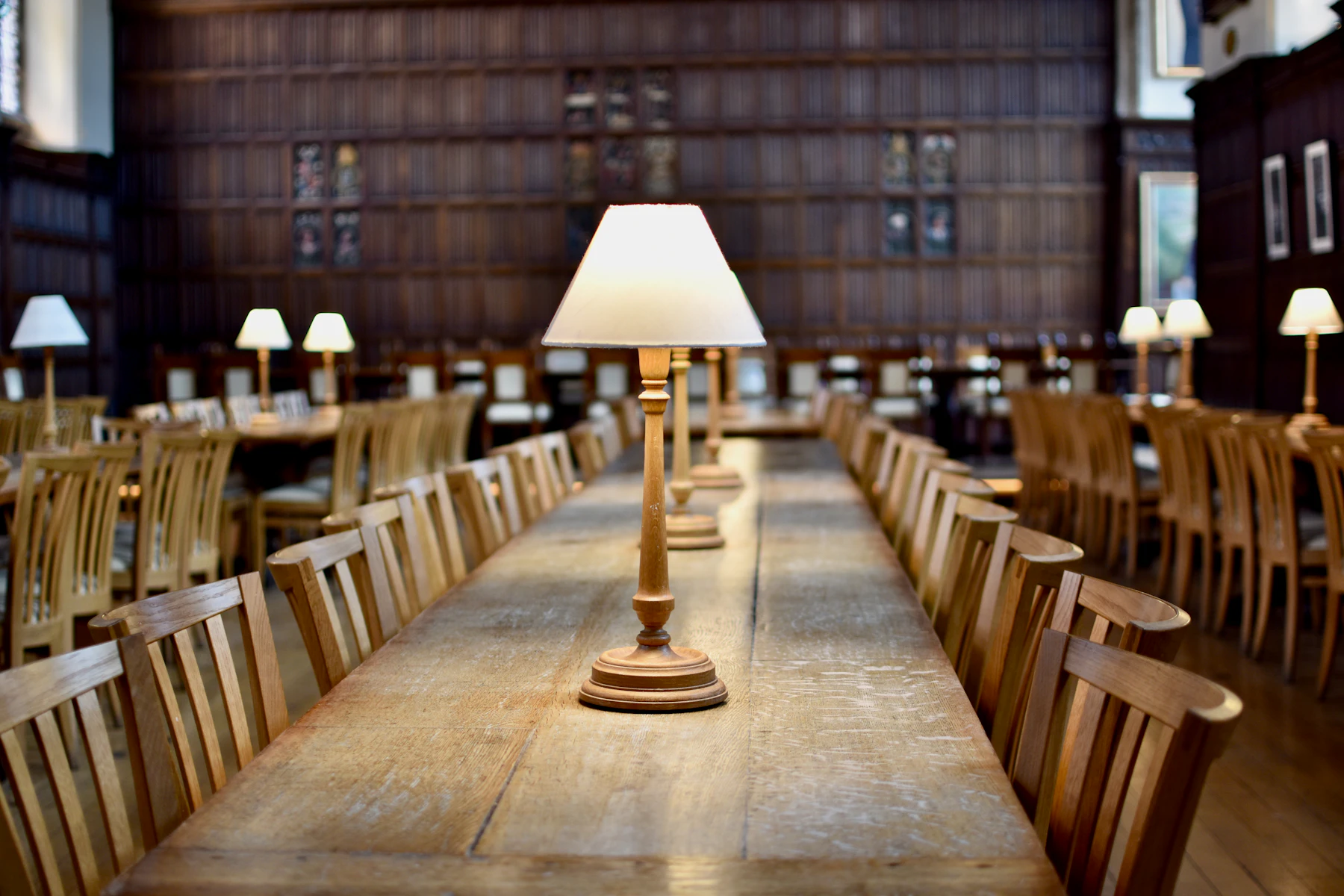A single brass reading lamp lit on a long oak table in a dim reading room