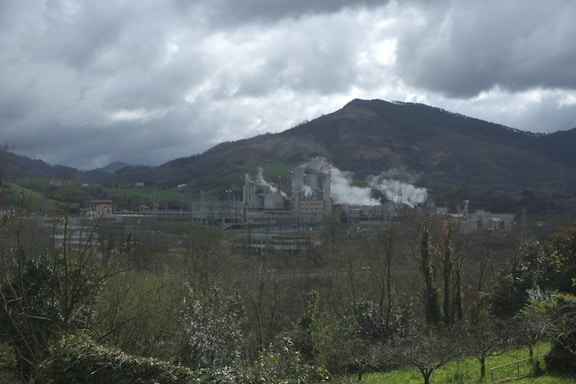An industrial facility is producing smoke amidst a scenic landscape featuring rolling hills and dense clouds. The facility is surrounded by greenery with a backdrop of mountains. The sky is overcast, adding to the moody atmosphere.