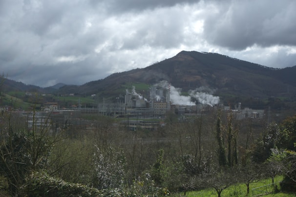 An industrial facility is producing smoke amidst a scenic landscape featuring rolling hills and dense clouds. The facility is surrounded by greenery with a backdrop of mountains. The sky is overcast, adding to the moody atmosphere.
