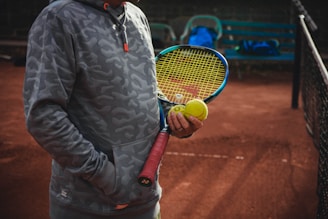 A person stands on a clay tennis court wearing a gray camouflage hoodie, holding a tennis racket with a yellow string pattern and a tennis ball. The person appears to be ready to play, with one hand in the pocket of the hoodie. In the background, there is a metal bench with a blue bag placed on it, and a partial view of the tennis net can be seen.