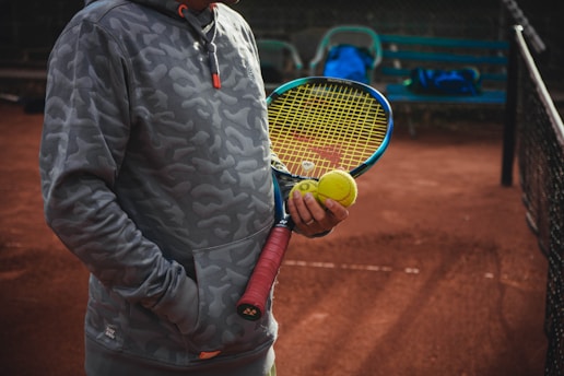 A person stands on a clay tennis court wearing a gray camouflage hoodie, holding a tennis racket with a yellow string pattern and a tennis ball. The person appears to be ready to play, with one hand in the pocket of the hoodie. In the background, there is a metal bench with a blue bag placed on it, and a partial view of the tennis net can be seen.