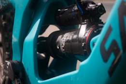 A close-up view of the suspension component of a bicycle, with a focus on the shock absorber. The frame and parts of the bicycle are painted in turquoise, with the shock absorber featuring a black and metallic design. The image captures the mechanical details and branding of the component.