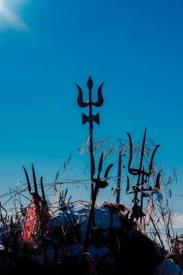 A group of tridents is set against a clear blue sky, with vegetation and cloth pieces intertwined around the handles. The central trident stands prominently, silhouetted elegantly while sunlight illuminates the scene.