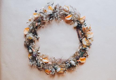A circular wreath made of dried flowers and foliage, featuring a mix of creamy whites, soft oranges, and subtle greens. The arrangement includes petals, leaves, and delicate sprigs, creating a rustic and organic aesthetic against a neutral background.