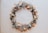 A circular wreath made of dried flowers and foliage, featuring a mix of creamy whites, soft oranges, and subtle greens. The arrangement includes petals, leaves, and delicate sprigs, creating a rustic and organic aesthetic against a neutral background.