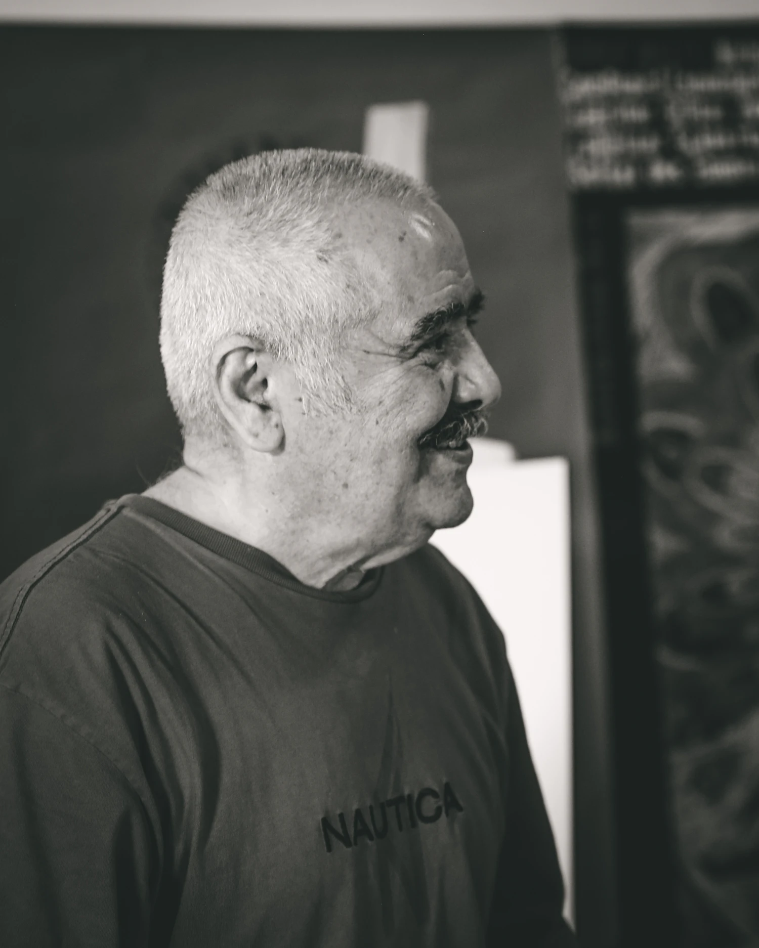 Black and white portrait of Ueli Aebersold, a grey-haired man with a moustache, smiling in front of a painting