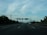 A highway stretches into the distance with several vehicles traveling on it. Overhead road signs are visible against a backdrop of scattered clouds and distant mountains. Dense green foliage lines the sides of the road, and a telecommunications tower is present on the left.