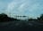 A highway stretches into the distance with several vehicles traveling on it. Overhead road signs are visible against a backdrop of scattered clouds and distant mountains. Dense green foliage lines the sides of the road, and a telecommunications tower is present on the left.