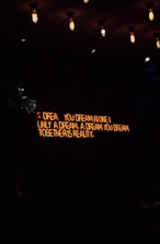 A dimly lit room features a neon sign with the words 'A dream you dream alone is only a dream. A dream you dream together is reality' against a dark brick wall. Several vintage-style light bulbs hang from the ceiling, casting a warm yellow glow.