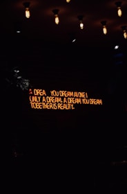 A dimly lit room features a neon sign with the words 'A dream you dream alone is only a dream. A dream you dream together is reality' against a dark brick wall. Several vintage-style light bulbs hang from the ceiling, casting a warm yellow glow.