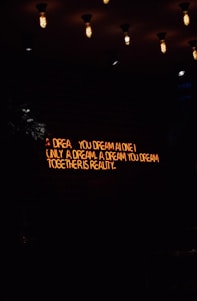 A dimly lit room features a neon sign with the words 'A dream you dream alone is only a dream. A dream you dream together is reality' against a dark brick wall. Several vintage-style light bulbs hang from the ceiling, casting a warm yellow glow.