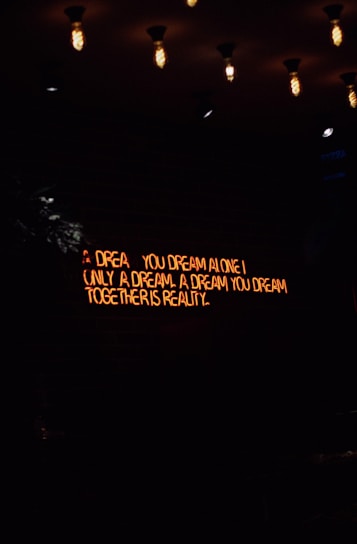 A dimly lit room features a neon sign with the words 'A dream you dream alone is only a dream. A dream you dream together is reality' against a dark brick wall. Several vintage-style light bulbs hang from the ceiling, casting a warm yellow glow.