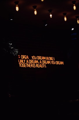 A dimly lit room features a neon sign with the words 'A dream you dream alone is only a dream. A dream you dream together is reality' against a dark brick wall. Several vintage-style light bulbs hang from the ceiling, casting a warm yellow glow.
