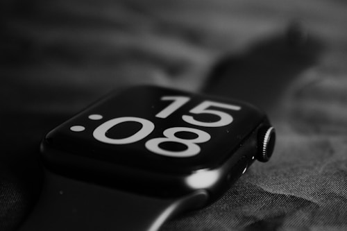 A modern digital watch with a prominent display showing the time 15:08. The watch has a sleek design, matte finish, and is photographed in black and white to highlight its minimalist aesthetic. The background is out of focus, drawing attention to the watch's details.
