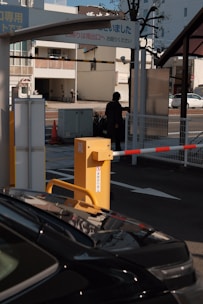A parking area with a barrier gate is visible, featuring a yellow gate arm and a sign with Japanese text. A black car is partially visible in the foreground, while a person in dark clothing stands near the barrier. The background includes a city street with buildings, a tree, and some traffic cones.