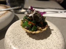A small tart filled with a colorful mixture of finely chopped vegetables, including green onions and carrots, topped with a dark spherical garnish and a small decorative flower. The tart rests on a speckled ceramic dish, with cutlery in the blurred background.