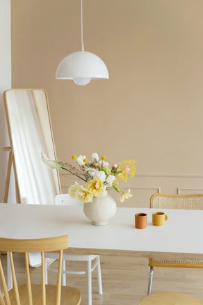 A minimalistic and modern interior scene featuring a light wooden dining table with a light-colored surface. On top of the table is a round white vase containing a diverse arrangement of flowers, including yellow roses and other colorful blossoms. Two ceramic mugs, one in a warm orange and the other in a muted brown, are placed beside the vase. Above the table hangs a contemporary white pendant lamp. In the background, a large floor mirror leans against a beige wall, complementing the neutral tones of the room. The setting includes two chairs, one made of natural wood and the other with a woven seat.