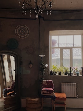 A cozy interior room with a rustic vibe, featuring a large window with several potted plants on the windowsill, allowing natural light to flood in. The walls are adorned with decorative circular wall hangings. A vintage chandelier hangs from the ceiling. Near the window, a chair with colorful fabrics is placed alongside a tall lamp and stacked wicker baskets.