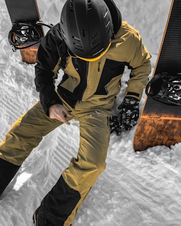A person wearing a black helmet and a yellow and black snowboarding outfit is seated on snow, surrounded by two snowboards. The snowboards have a wooden finish and bindings attached. The individual is adjusting their glove and appears to be resting or preparing for snowboarding.