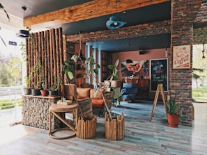 A cozy, rustic cafe interior with brick walls and wooden accents. There are various potted plants adding greenery, and comfortable seating arrangements including chairs and sofas. Coffee-themed decorations, including a sign and artwork, are displayed on the walls. Natural light fills the space through large windows, creating a warm and inviting atmosphere.
