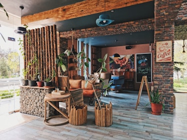 A cozy, rustic cafe interior with brick walls and wooden accents. There are various potted plants adding greenery, and comfortable seating arrangements including chairs and sofas. Coffee-themed decorations, including a sign and artwork, are displayed on the walls. Natural light fills the space through large windows, creating a warm and inviting atmosphere.