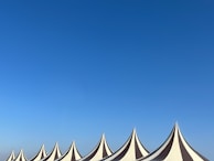 A series of pointed tent tops with alternating black and white stripes stand against a clear blue sky. The tents are arranged in a row, creating a rhythmic pattern across the bottom of the image.