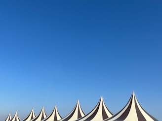 A series of pointed tent tops with alternating black and white stripes stand against a clear blue sky. The tents are arranged in a row, creating a rhythmic pattern across the bottom of the image.