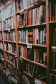 A cozy bookstore aisle lined with tall wooden bookshelves filled with a variety of hardcover books. The books are arranged in an orderly manner, and there are handwritten signs on the shelves. The overall ambiance suggests a warm, inviting space for book lovers.