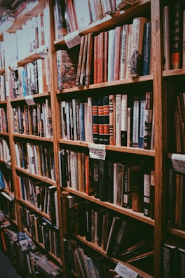 A cozy bookstore aisle lined with tall wooden bookshelves filled with a variety of hardcover books. The books are arranged in an orderly manner, and there are handwritten signs on the shelves. The overall ambiance suggests a warm, inviting space for book lovers.