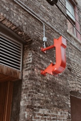 A brick wall of an urban building with a red, glossy letter 'J' sign mounted on it. Visible are the windowpanes, a set of metal pipes running horizontally, and a section of wooden paneling under an air vent.