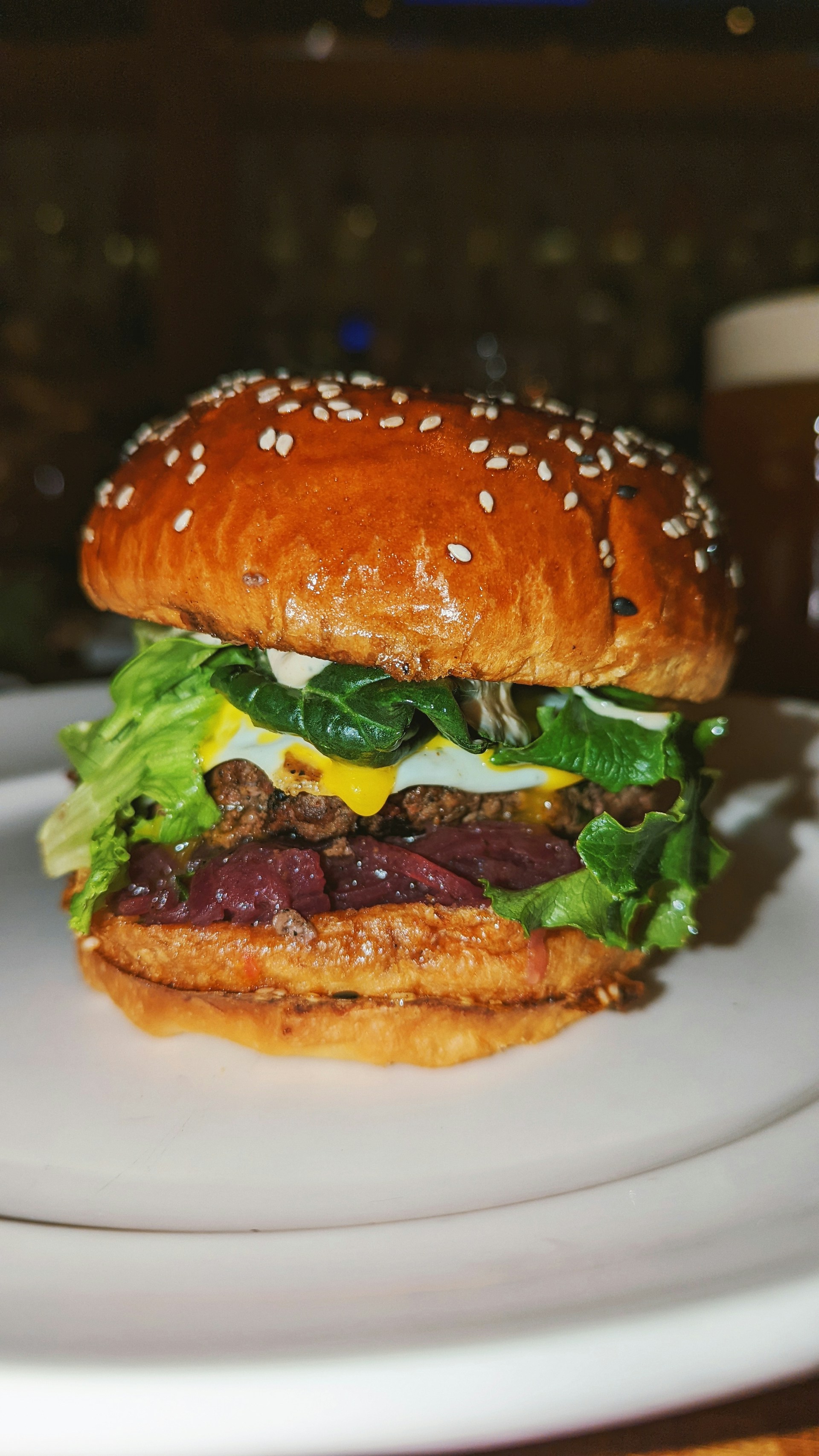 A juicy burger with a sesame seed bun, containing fresh lettuce, a patty, cheese, and other toppings. The burger is placed on a white plate, with a blurred background suggesting a restaurant setting.