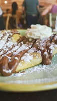 A close-up view of a dessert featuring a plate of waffles drizzled with chocolate sauce and dusted with powdered sugar. There is a dollop of whipped cream on the side. In the background, a blurry interior setting with people seated and wooden furniture.