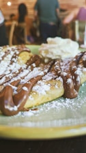 A close-up view of a dessert featuring a plate of waffles drizzled with chocolate sauce and dusted with powdered sugar. There is a dollop of whipped cream on the side. In the background, a blurry interior setting with people seated and wooden furniture.