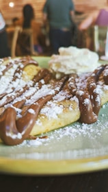 A close-up view of a dessert featuring a plate of waffles drizzled with chocolate sauce and dusted with powdered sugar. There is a dollop of whipped cream on the side. In the background, a blurry interior setting with people seated and wooden furniture.
